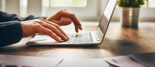 A close-up view of hands typing on a laptop keyboard, placed on a wooden desk. The background features soft, natural light filtering through windows, enhancing a focused work atmosphere.