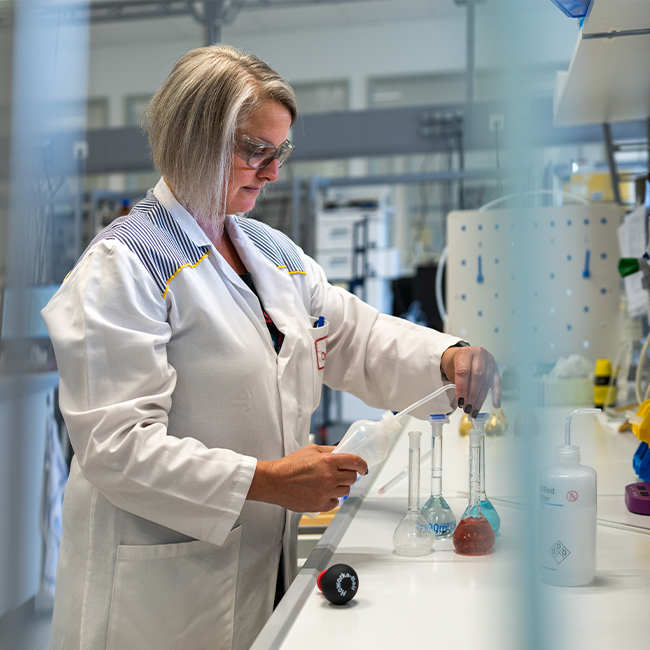 A scientist wearing a white lab coat and glasses is working in a laboratory. They are using a pipette to transfer liquids between various glass flasks, with lab equipment and materials visible in the background.