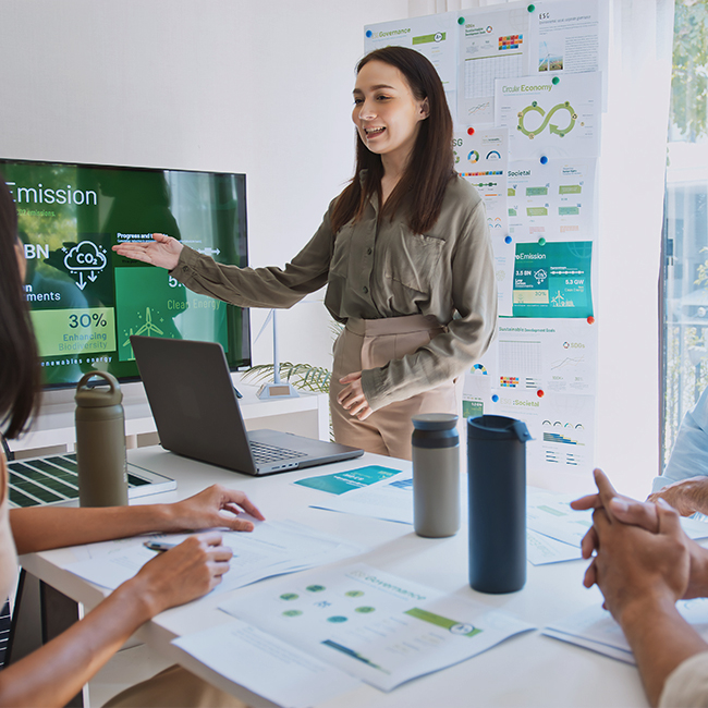 A woman stands in front of a screen displaying emission data during a presentation. She gestures towards the screen, while seated individuals at a table, with laptops and water bottles, listen attentively. Charts and graphics about sustainability and emissions are visible in the background.